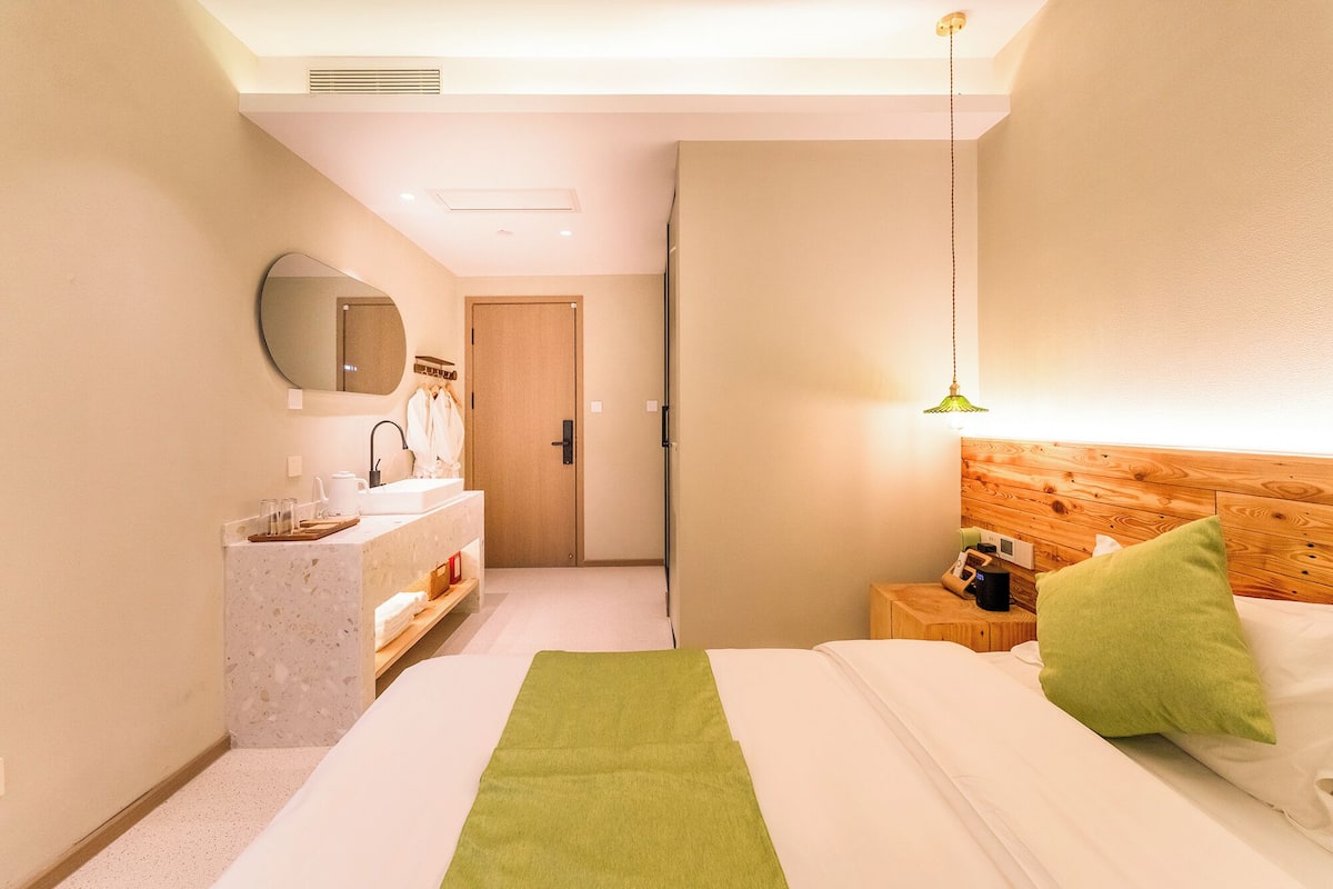 A hotel room with a bed, a wooden headboard, a nightstand, a bedside table, a mirror, a sink, and a green rug.