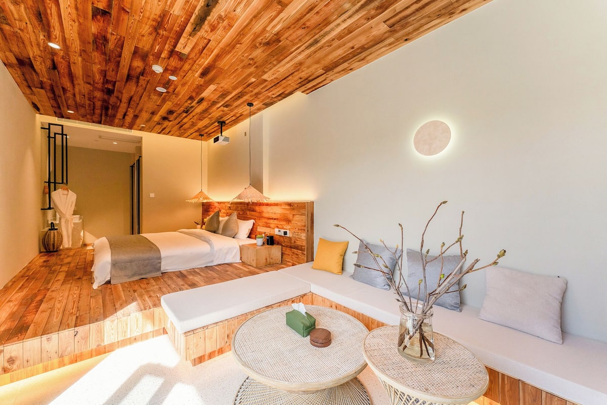 A modern hotel room with a wooden ceiling, a sofa, a bed, and a round table with a vase.