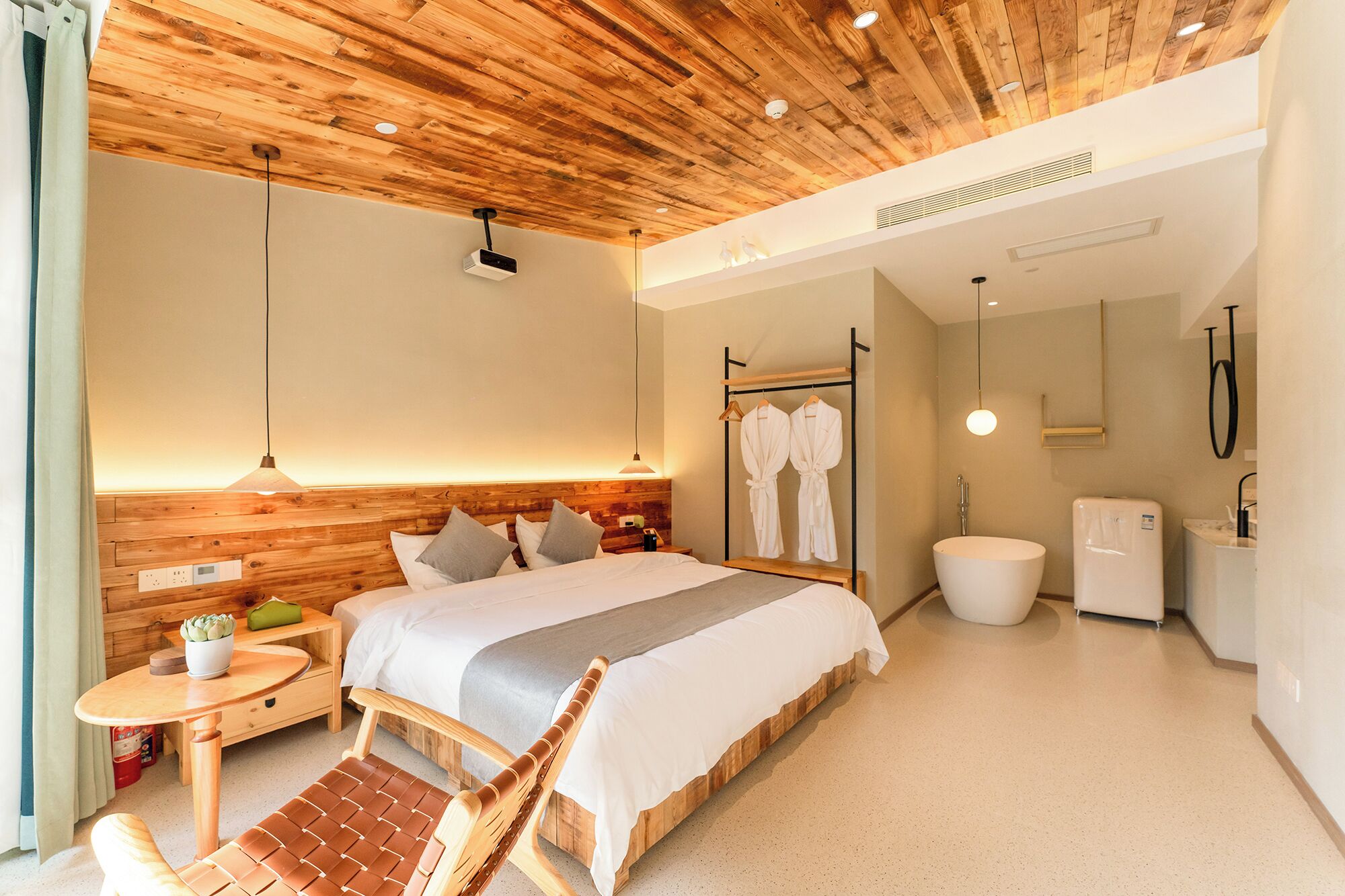 A modern hotel room with a wooden ceiling, a bed with a grey blanket, a bedside table with a plant, a chair, and a bathroom with a white sink and mirror.