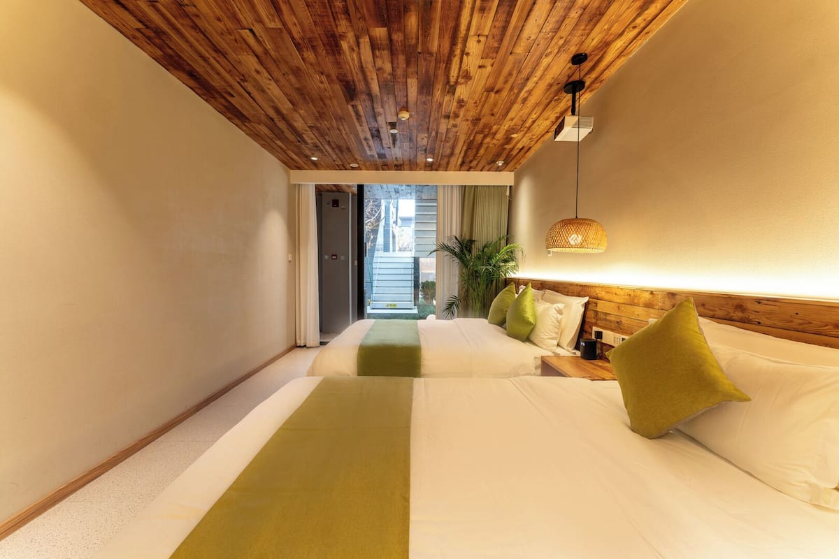 A hotel room with a wooden ceiling, a bed with white bedding and green pillows, a bedside table with a lamp, and a view of the outside.