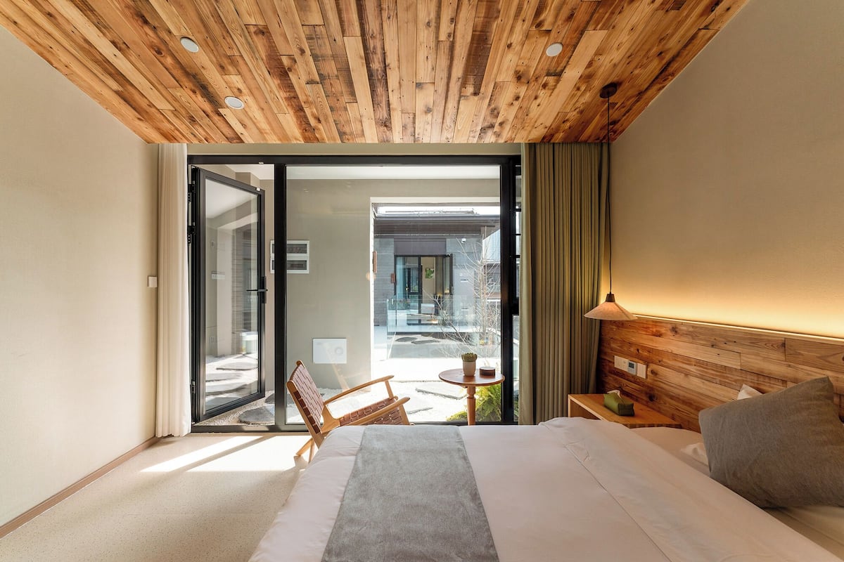 A hotel room with a wooden ceiling, a bed, a chair, a table, and a view of the outside.