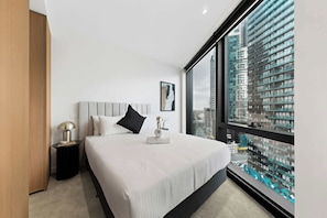 Ein modernes Hotelzimmer mit einem großen Bett, einem Nachttisch, einer Nachttischlampe, einem gerahmten Bild und Blick auf die Stadt durch ein großes Fenster.