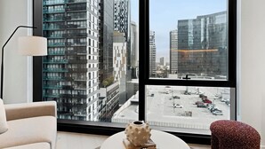 Moderni hotellihuone, jossa on suuri ikkuna, josta avautuu näkymä korkeisiin rakennuksiin ja parkkipaikalle.