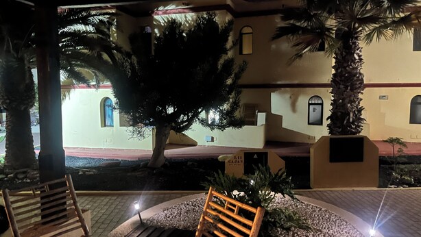 Un espace extérieur aménagé avec des chaises et une table en bois, entouré d’un jardin planté de palmiers et, au fond, d’un bâtiment doté de fenêtres en arc de cercle.