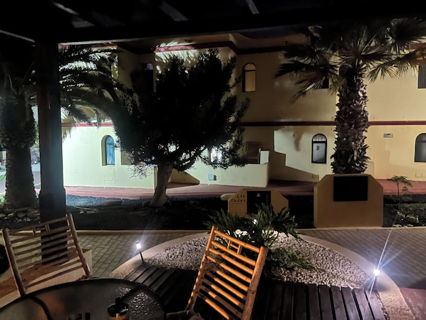Ein Außenbereich mit Holzkästen und einem Tisch, umgeben von einem Garten mit Palmen und einem Gebäude mit Rundbogenfenstern im Hintergrund.