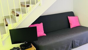 Une pièce avec un canapé gris, deux coussins roses, un téléviseur à écran plat, une table d’appoint en bois et un escalier avec des rampes blanches.