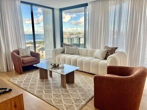 Ein modernes Wohnzimmer mit Sofa, Sessel und Couchtisch, großen Fenstern mit Meerblick und einem gemusterten Teppich.