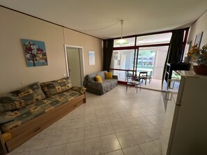 Ein Wohnzimmer mit einer Couch, einem Esstisch mit Stühlen, einem Kühlschrank und einem großen Fenster mit Vorhängen.