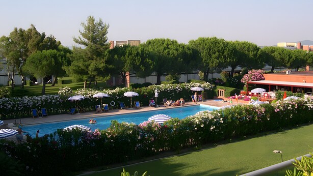 Area piscina con lettini e ombrelloni, immersa nel verde con edifici sullo sfondo.