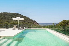 A clear swimming pool with a glass barrier, lounge chairs, and an umbrella, set against a backdrop of a hillside and the sea.