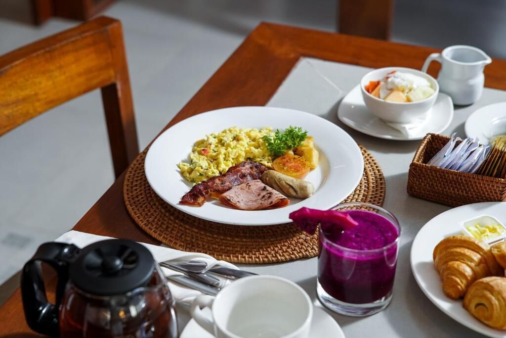 Une table dressée pour le petit-déjeuner avec des œufs brouillés, du bacon, du jambon, des toasts, un bol de soupe, des croissants et un verre d’une boisson violette.