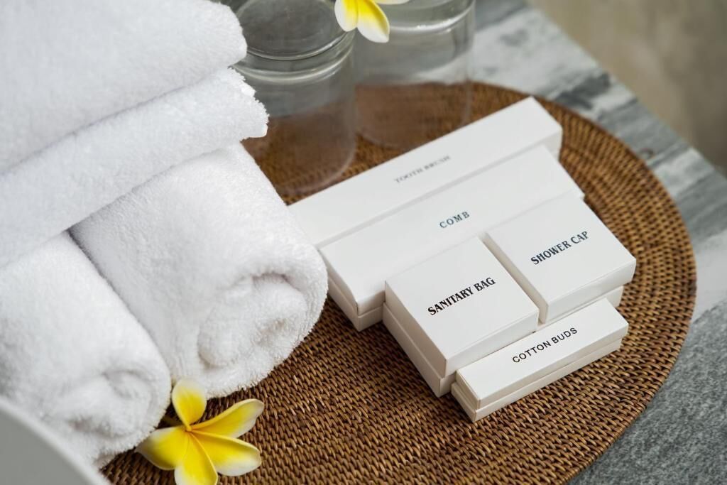 Des serviettes blanches soigneusement pliées, un plateau en osier et des boîtes d’articles de toilette étiquetées, contenant notamment un bonnet de douche, un sac d’hygiène et des cotons-tiges.