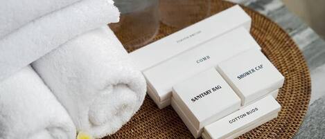 Des serviettes blanches soigneusement pliées, un plateau en osier et des boîtes d’articles de toilette étiquetées, contenant notamment un bonnet de douche, un sac d’hygiène et des cotons-tiges.