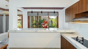 Una cocina moderna con encimera blanca, armarios de madera, luces colgantes y vistas al exterior con zonas verdes.