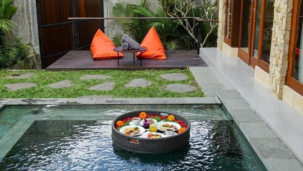 Una piscina con un espacio de comedor flotante, una zona de estar con cojines naranja y un sendero de piedra.