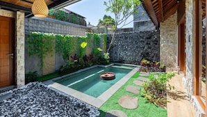 Una piscina con un hogar de fuego, rodeada por un muro de piedra, follaje y una pérgola de madera.