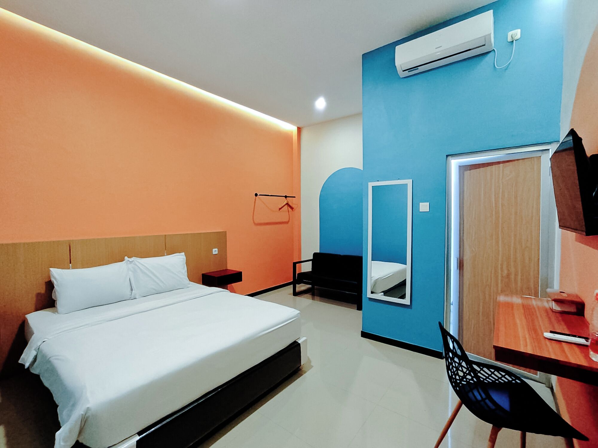 A hotel room with a bed, a desk, a chair, a mirror, and an air conditioning unit.