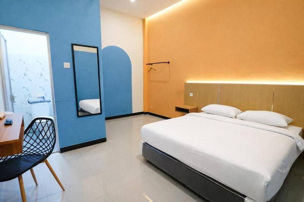 Una habitación de hotel con cama, escritorio, silla, espejo y lámpara de pared.