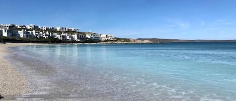 Una playa costera con agua cristalina, orilla de piedras y un complejo de edificios blancos al fondo.