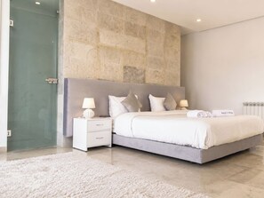 A modern bedroom with a large bed, a nightstand, a glass shower enclosure, and a stone wall. - Luxurious Villa in Gammarth with Private Pool and Garden Views (Gammarth)