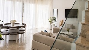 Ein modernes Wohnzimmer mit einer Glastreppe, einem Essbereich mit runden Tischen und einem großen Fenster mit transparenten Vorhängen.