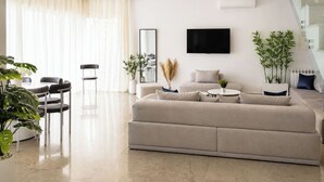 Ein modernes Wohnzimmer mit einer Couch, einem Esstisch mit Stühlen, einem an der Wand befestigten Fernseher und Topfpflanzen.
