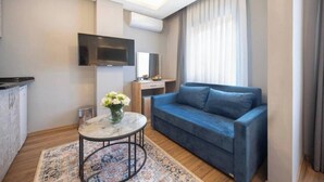 Ein modernes Wohnzimmer mit einem blauen Sofa, einem kleinen runden Tisch mit Blumenvase, einem flachbildfernseher an der Wand und einem großen Fenster mit Vorhängen.