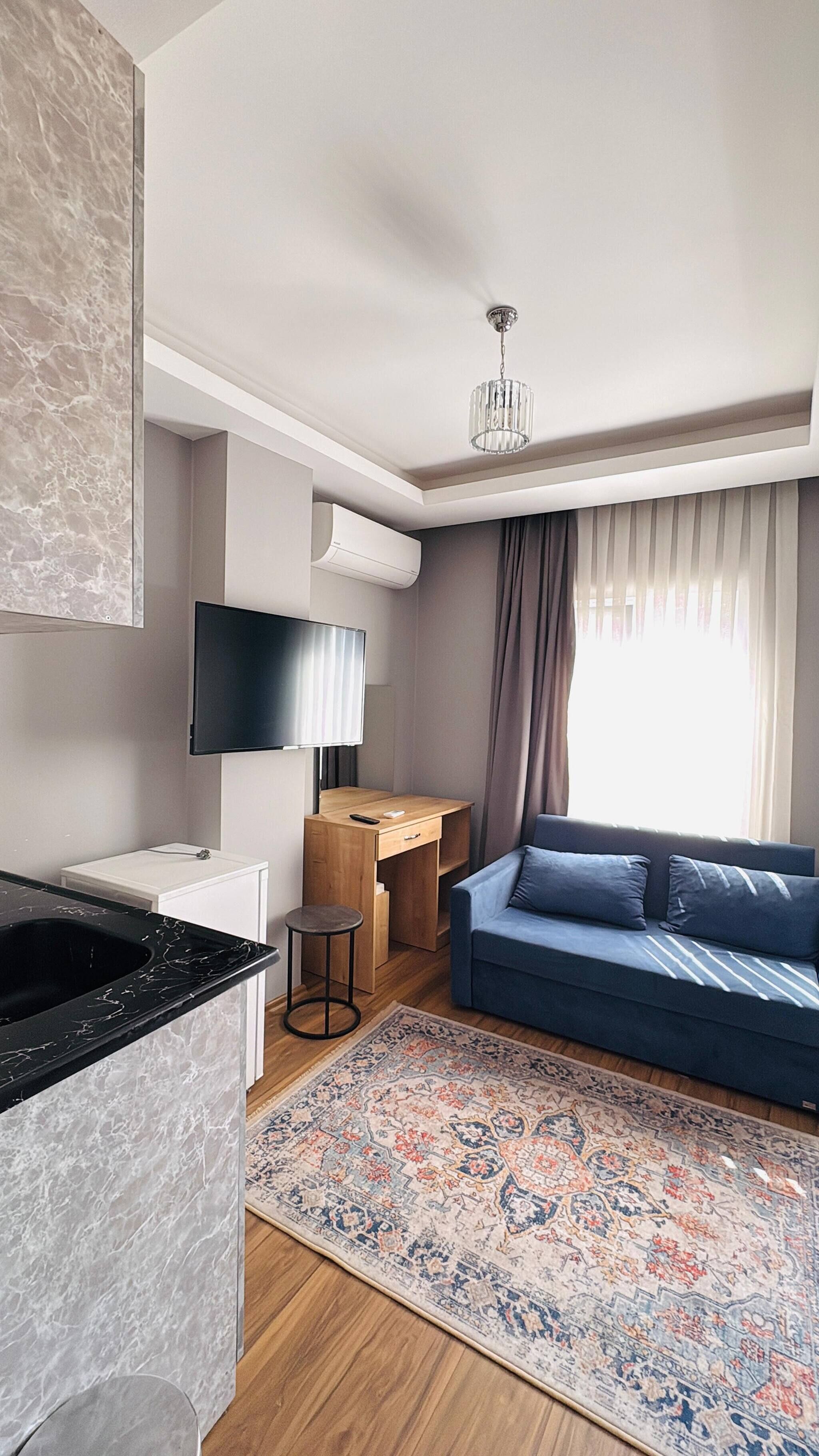Een moderne woonkamer met een blauwe bank, een televisie, een houten bureau en een keuken met een marmeren aanrecht.