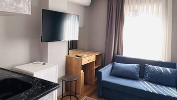 Een moderne woonkamer met een blauwe bank, een televisie, een houten bureau en een keuken met een marmeren aanrecht.