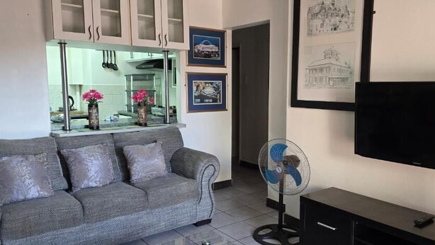 A living room with a grey sofa, a TV, a fan, and framed pictures on the wall. - Summer sands Air b n b (Durban)