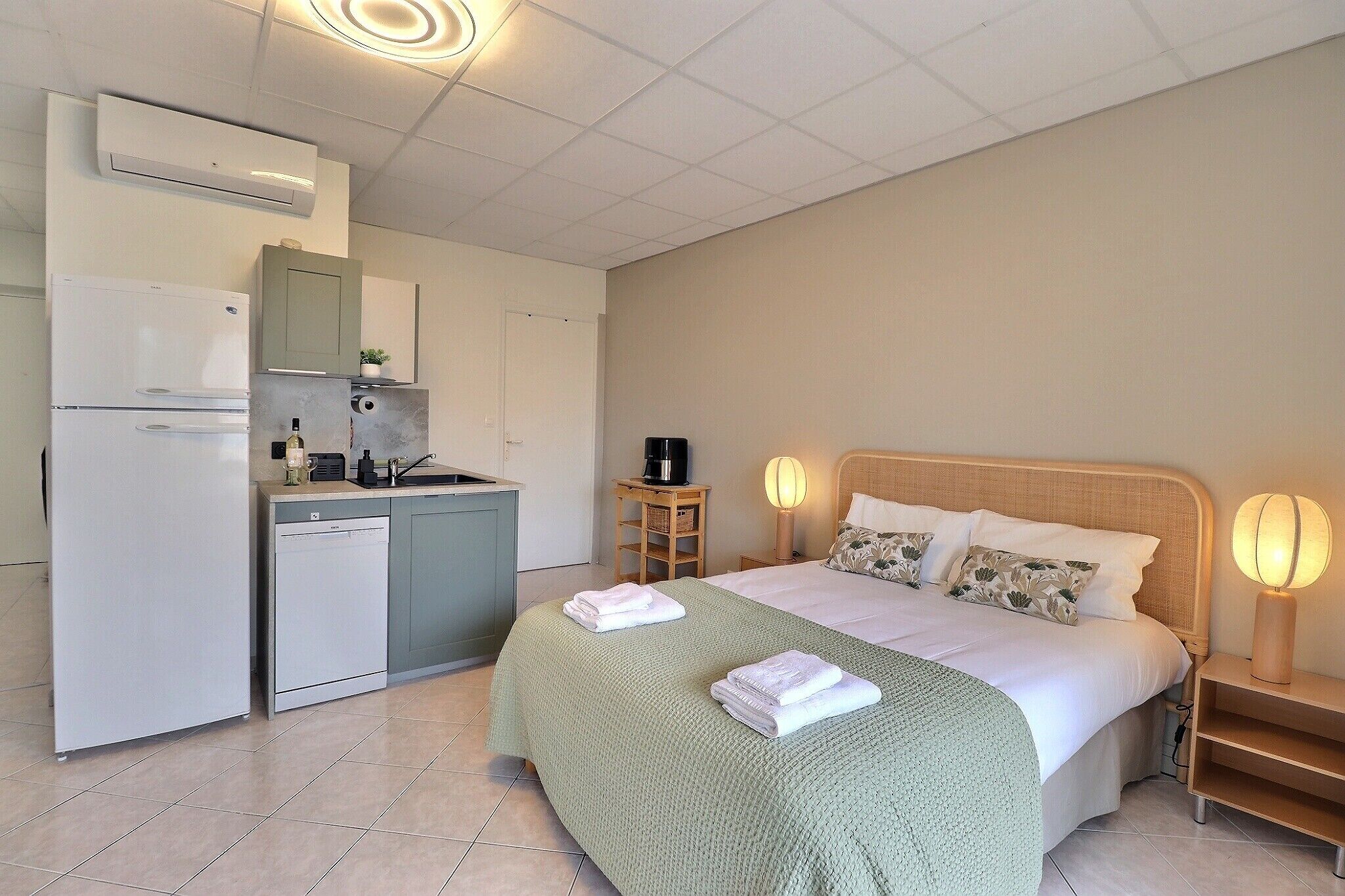 A compact hotel room with a kitchenette, a bed with two pillows, a bedside table with a lamp, and a small side table with a coffee machine.