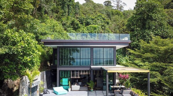 Ein modernes, zweistöckiges Haus mit großen Fenstern, einer Terrasse mit Gartenmöbeln und üppigem Grün drumherum.