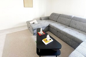 A modern living room with a grey sectional sofa, a black coffee table with magazines, and a small ottoman with a white towel.
