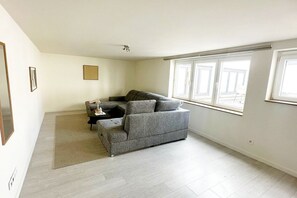Ein Wohnzimmer mit grauem Sofa, einem kleinen Tisch und einem Teppich. Zwei Fenster mit Jalousien, und die Wände sind weiß.