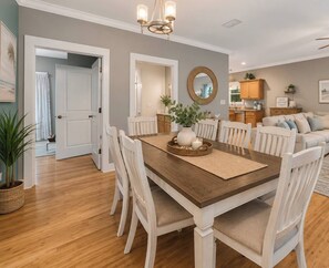 A dining area with a wooden table, white chairs, a chandelier, and a kitchen in the background. - 5003 R3 Serene Beach Cottage in Gulf Shores 3BR 2 Bath, Sleeps 10 (Gulf Shores)