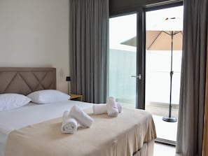 Ein Hotelzimmer mit einem Bett, Kissen, einem Nachttisch mit Lampe und Blick durch eine Glastür auf einen Außenbereich mit Sonnenschirm.