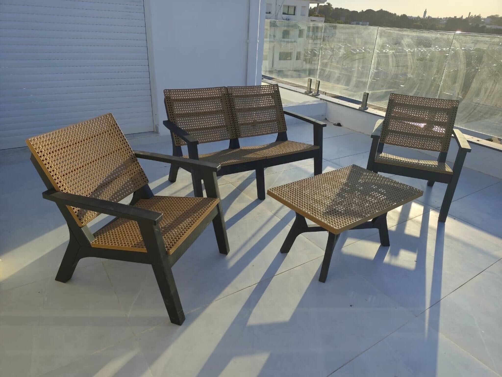 A balcony with four wicker chairs and a small table, a white wall, and a view of a cityscape.