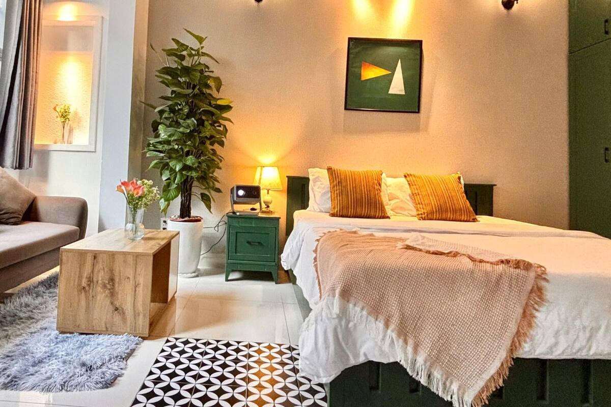 Quarto de hotel com cama, mesa de cabeceira, abajur, planta em vaso e uma obra de arte pendurada na parede.