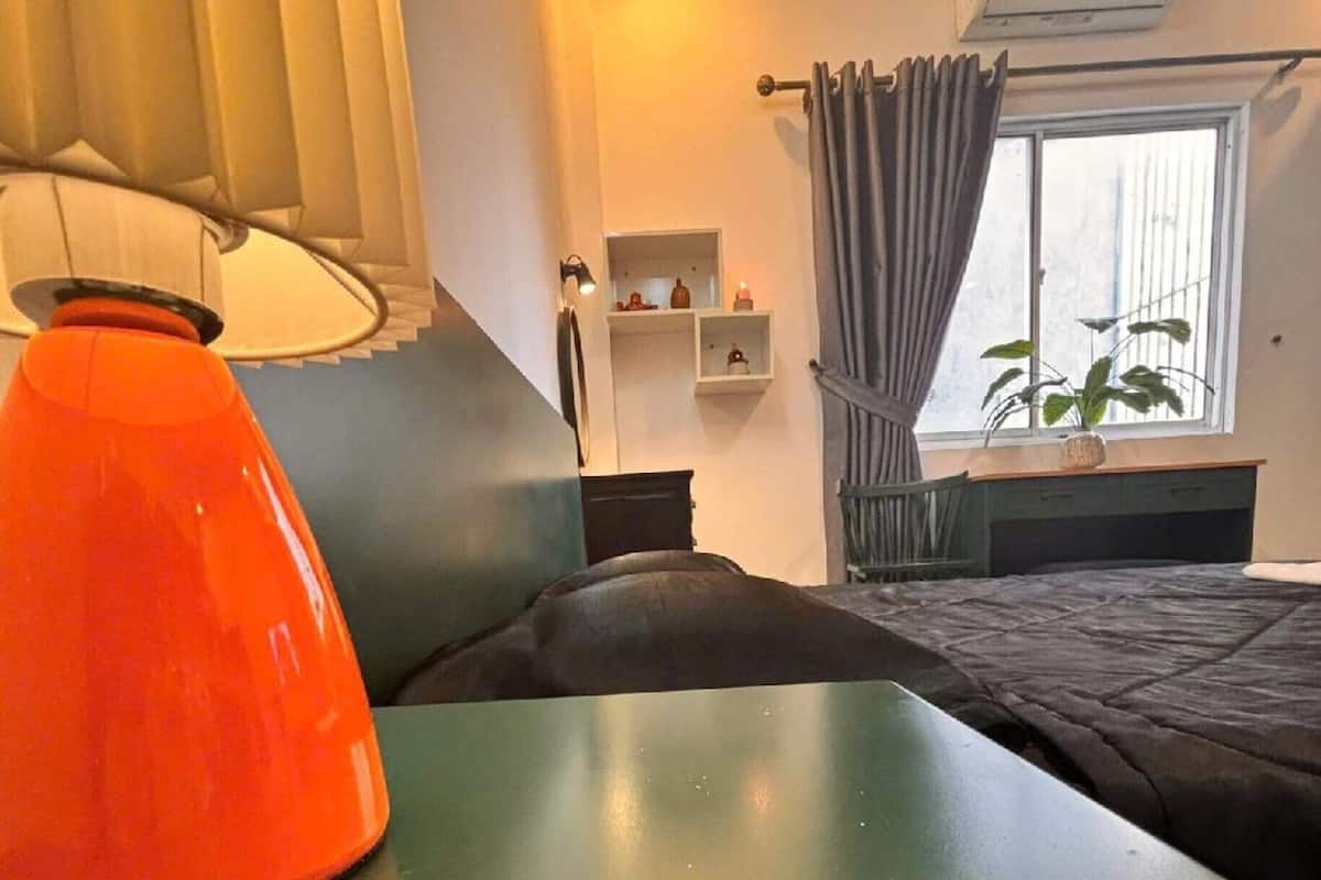 Quarto com cama, mesa com luminária, janela com cortinas, prateleira com objetos decorativos e ar condicionado.