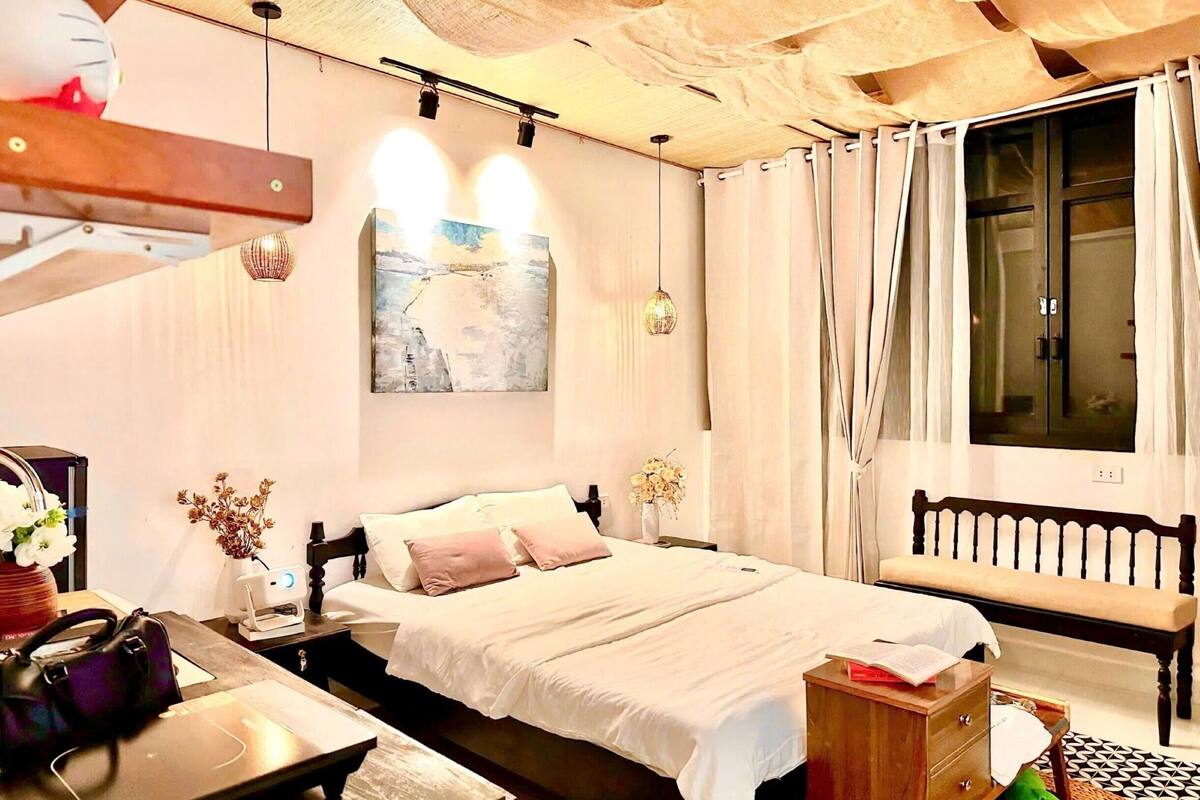 Quarto com cama, mesa de cabeceira, luminária, obra de arte, janela com cortinas e um banco.