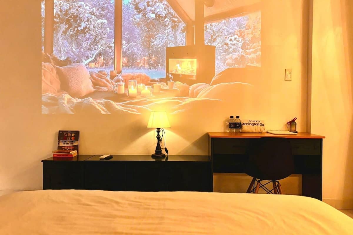 Quarto de hotel com cama, escrivaninha com luminária, cadeira, tela de projetor exibindo uma cena da natureza e uma pequena mesa com uma garrafa de água e um travesseiro.