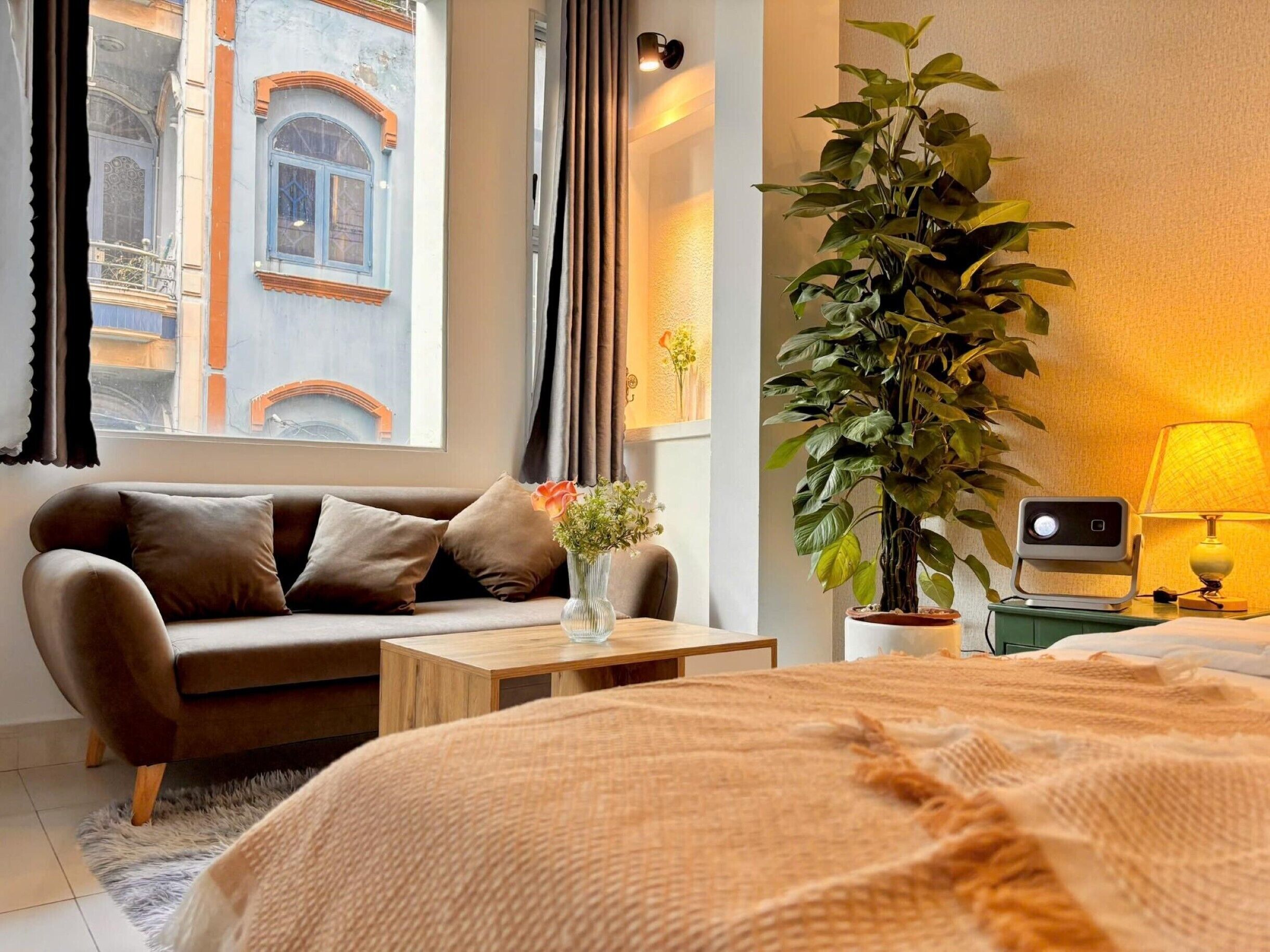 Una camera d'albergo con un letto, un divano, un tavolino da caffè, una pianta in vaso di grandi dimensioni, una lampada e vista su un edificio.