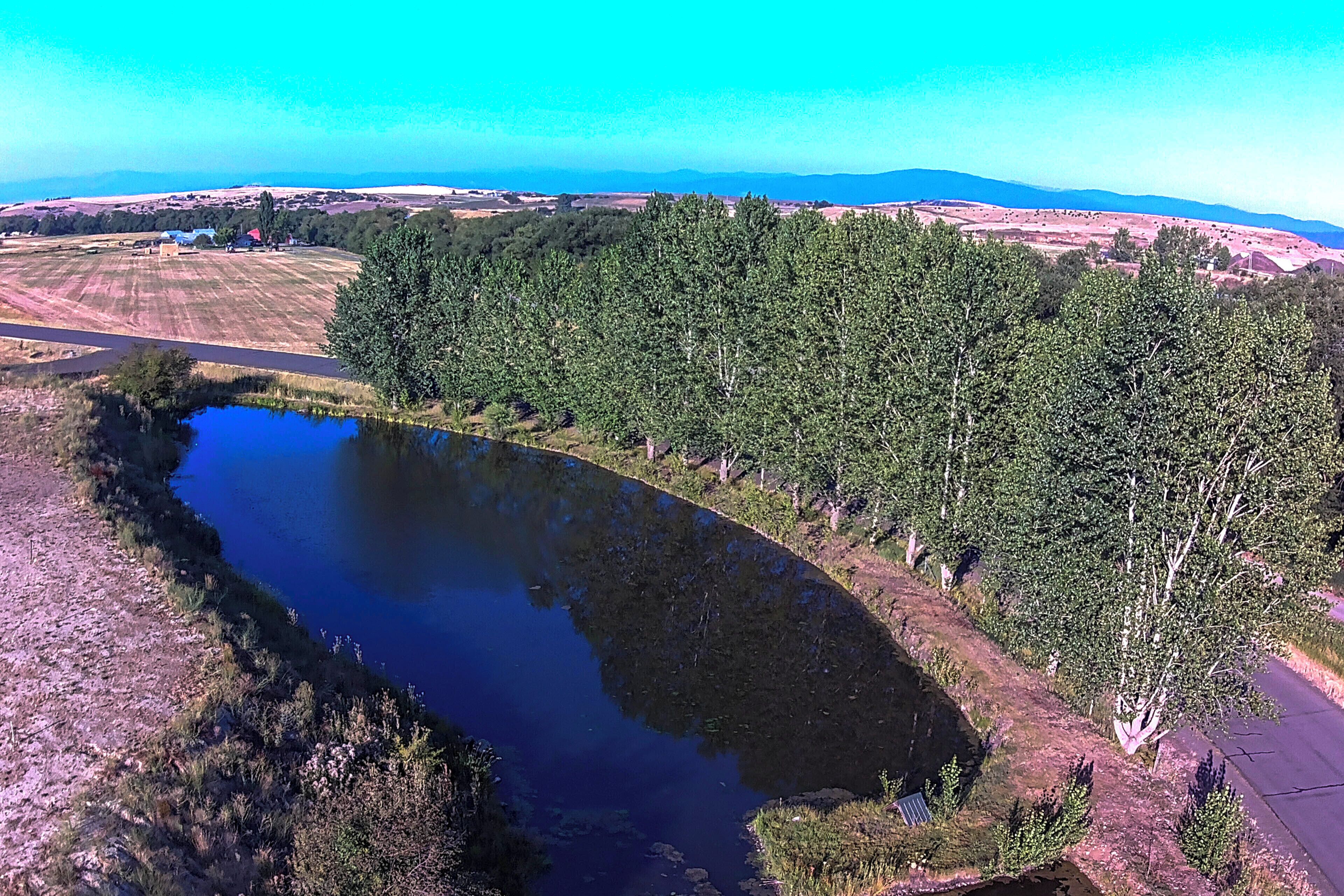 Une vaste étendue d’eau entourée d’une forêt dense, avec en arrière-plan un paysage de champs et de quelques bâtiments.