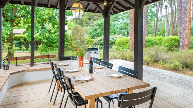 Une terrasse extérieure couverte, avec une table en bois dressée pour les repas, entourée de chaises, et un barbecue à l’arrière-plan.