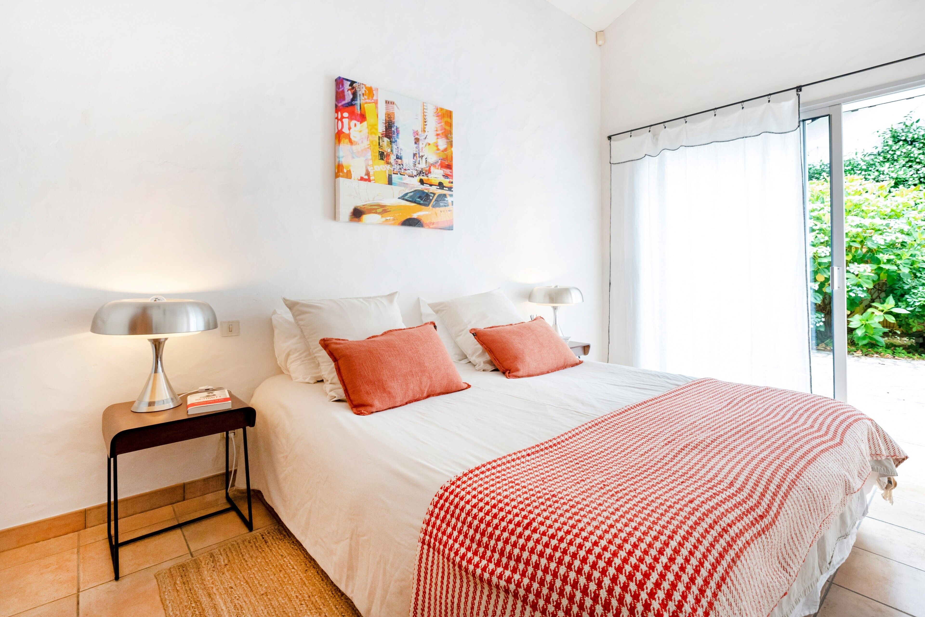 Une chambre à coucher avec un lit, des oreillers, une lampe, un tableau et une vue sur la verdure.