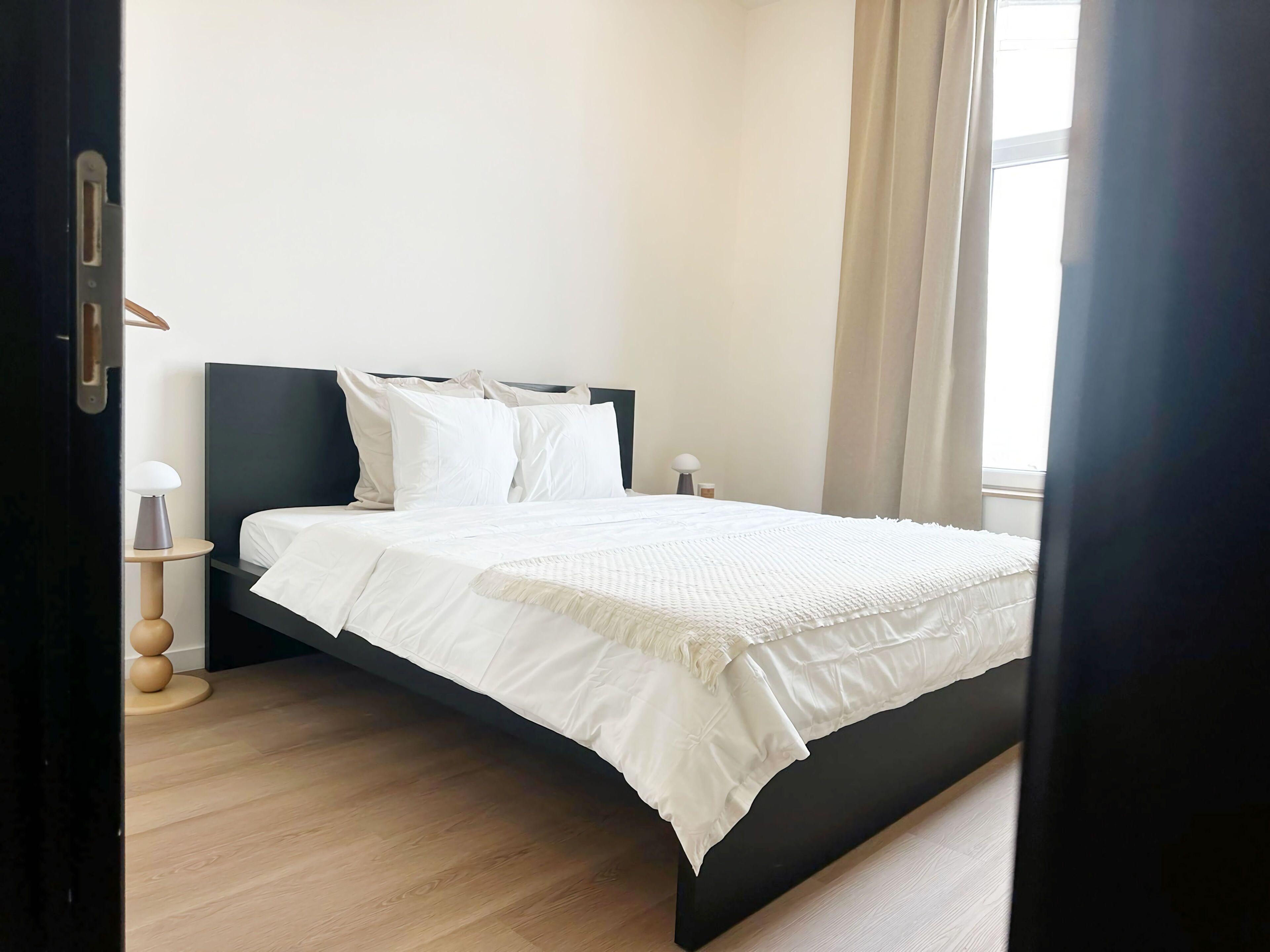 Een moderne slaapkamer met een bed, nachtkastje, lamp en een raam met gordijnen.