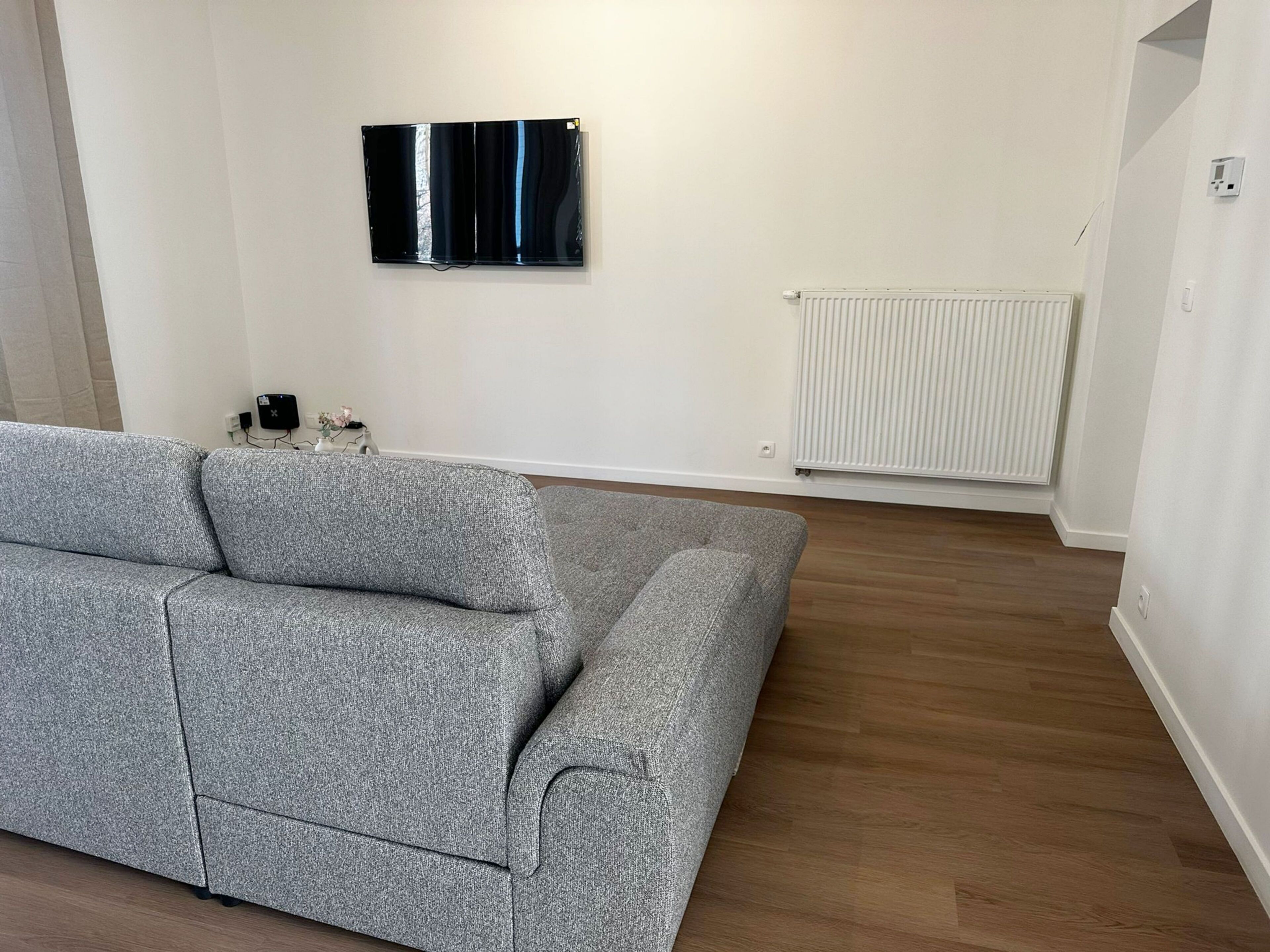 Een woonkamer met een grijze bank, een flatscreen televisie aan de muur en een witte radiator.
