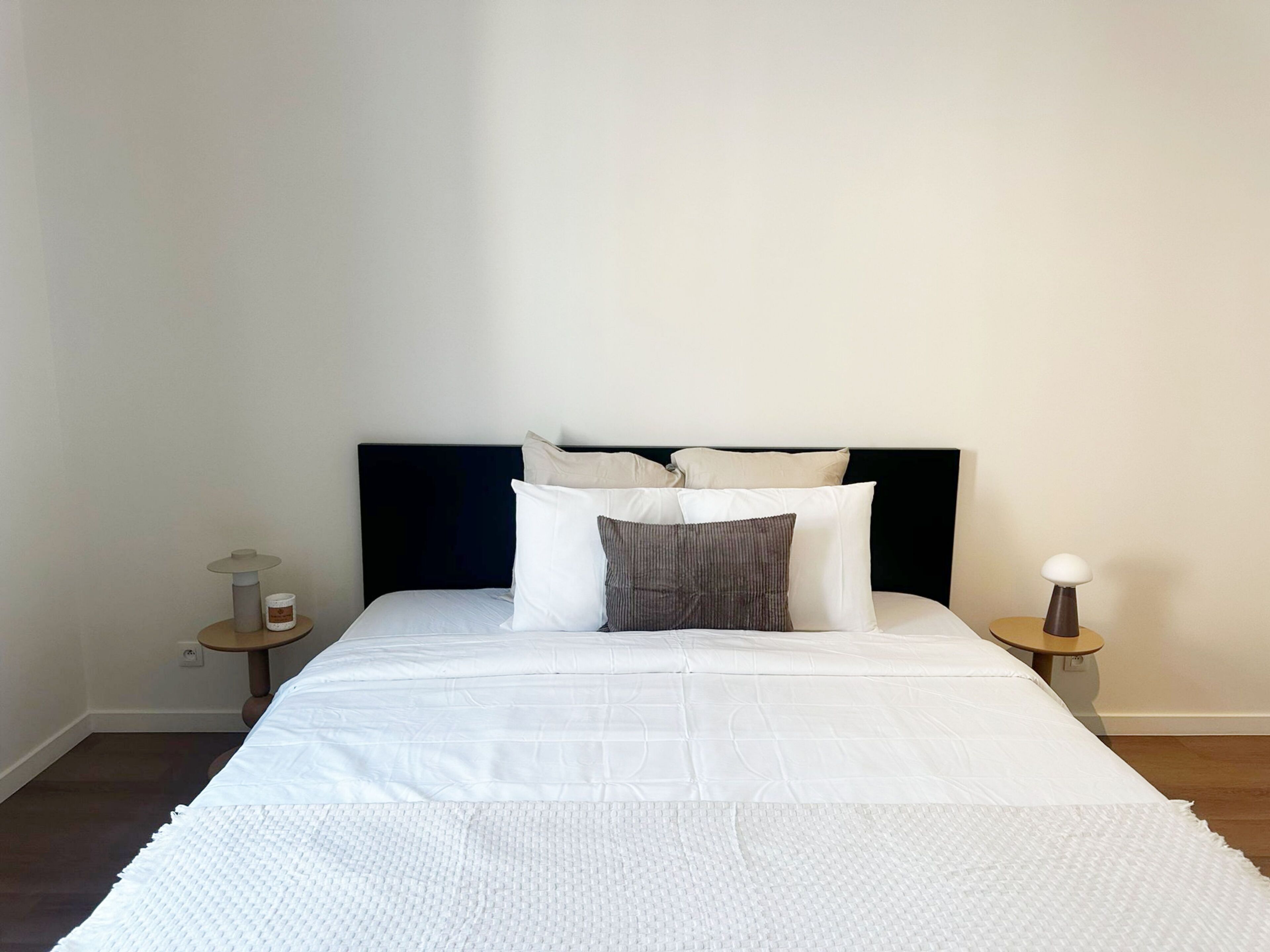 Een net opgemaakt bed met een donker hoofdbord, witte beddengoed en een paar kussens. Er zijn twee nachtkastjes met lampen en een klein tafeltje met een kaars. De kamer heeft effen muren en een houten vloer.