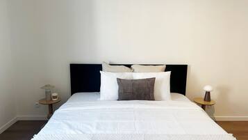 Een net opgemaakt bed met een donker hoofdbord, witte beddengoed en een paar kussens. Er zijn twee nachtkastjes met lampen en een klein tafeltje met een kaars. De kamer heeft effen muren en een houten vloer.