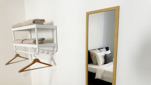 Estante blanco montado en la pared con toallas y perchas de madera, un espejo grande con marco de madera, y una cama con ropa de cama y almohadas blancas.
